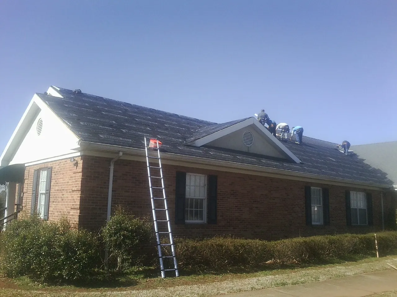 Crew tearing shingles off a brick ranch roof with a ladder set — Emergency Roof Repair work by our crew in West Hempstead