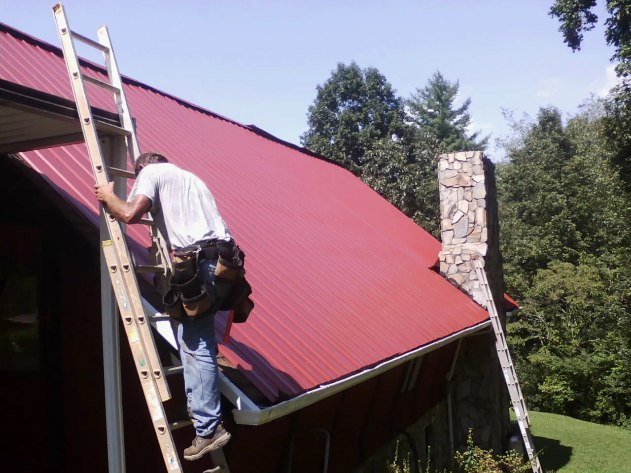Roofer on a ladder beside a red metal roof and stone chimney — Soffit & Fascia Repair by Roofing Brothers in West Hempstead
